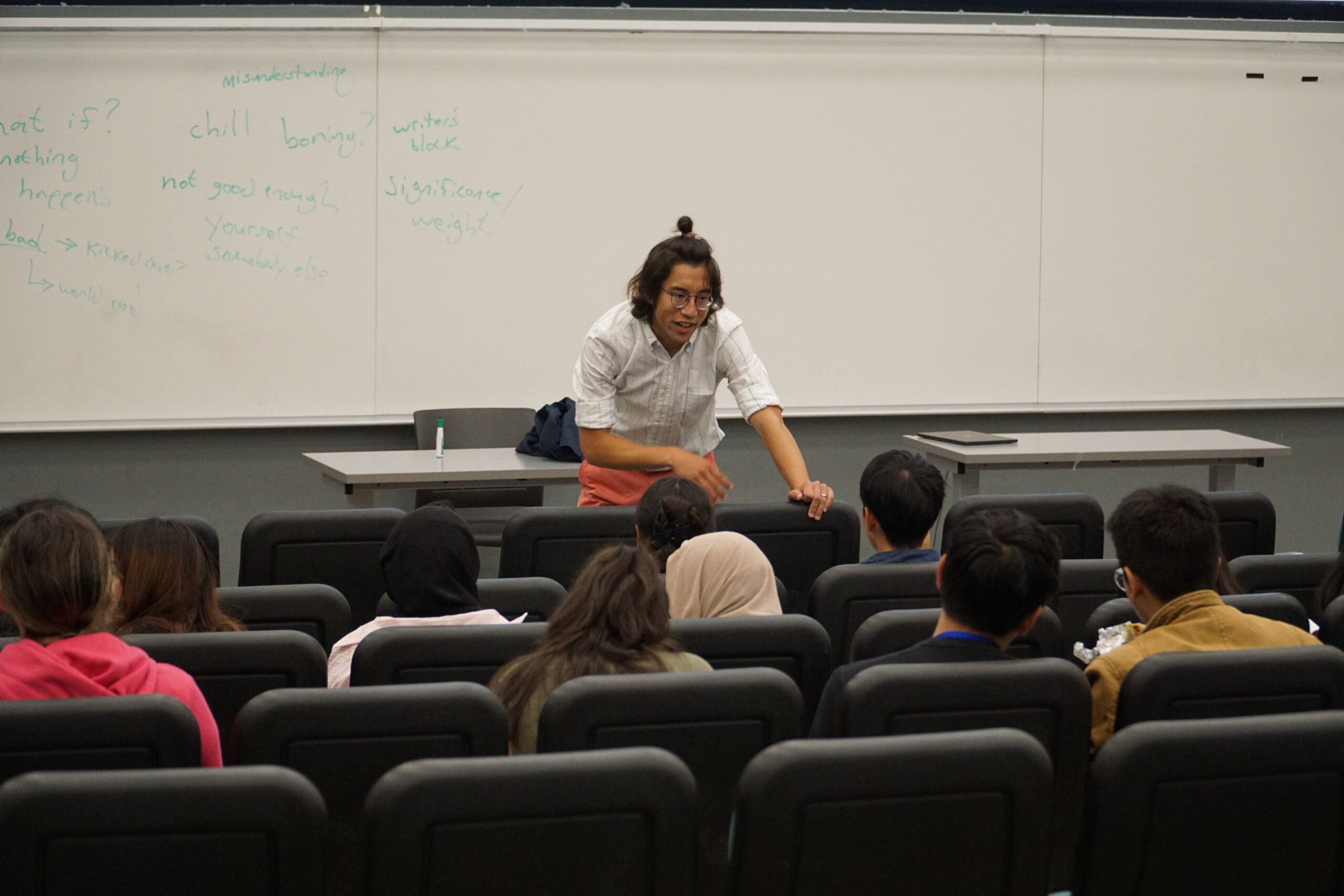 teacher at front of classroom, leaning toward seated students and smiling