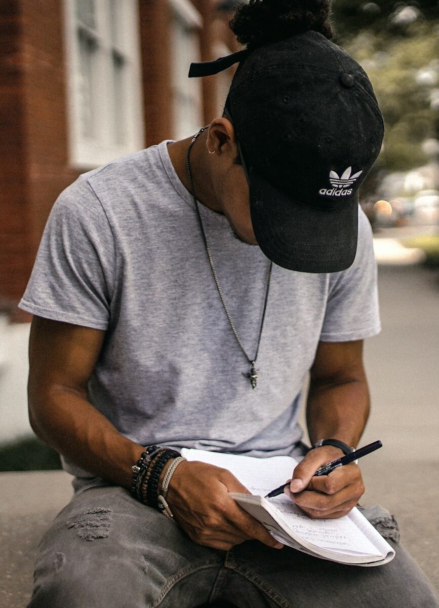 person in black adidas cap sitting on bench writing on notebook