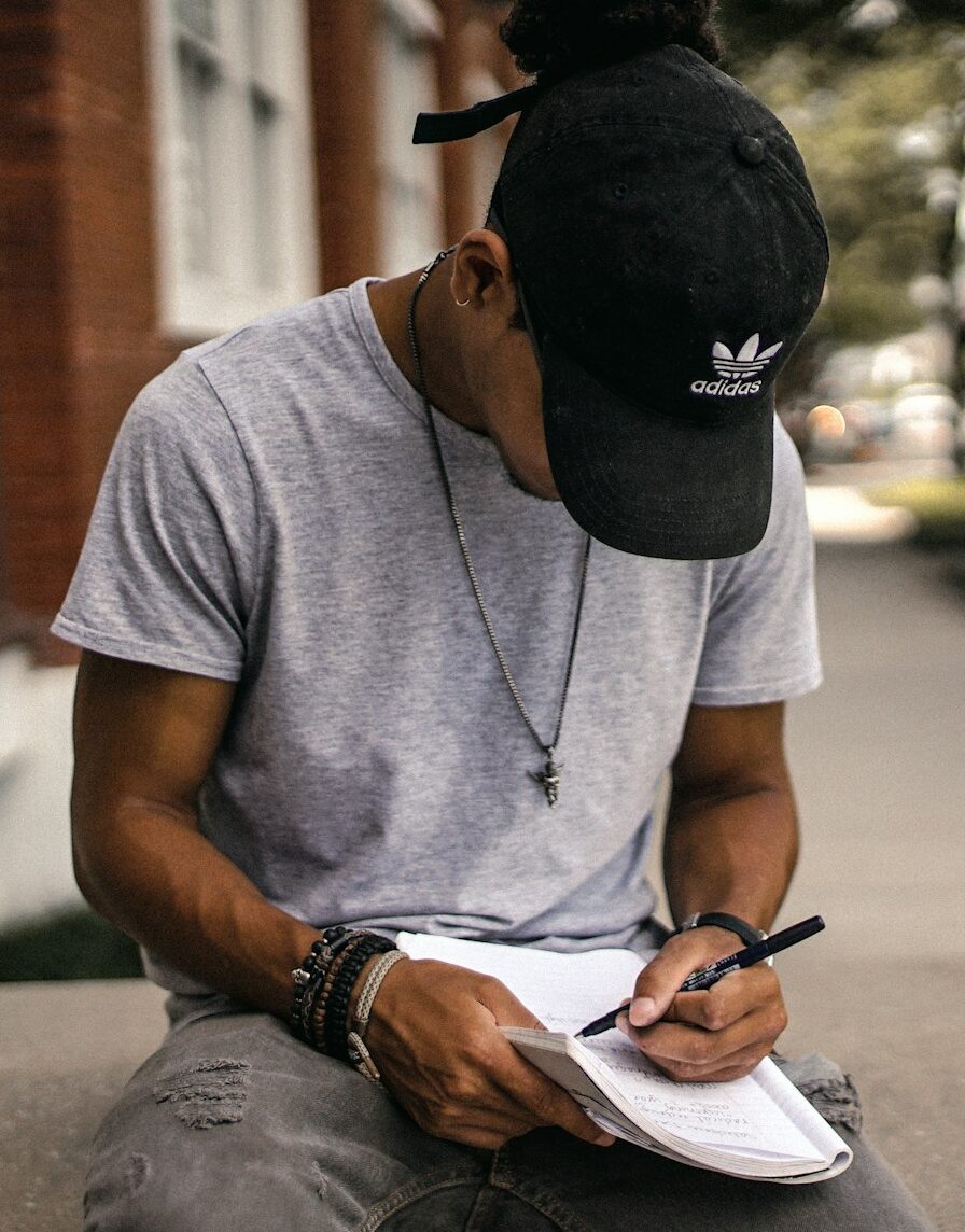 person in black adidas cap sitting on bench writing on notebook